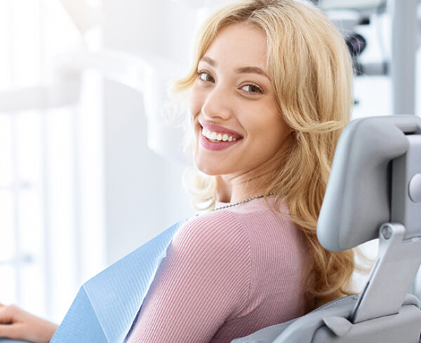 Smiling and relaxed young woman sitting at dental chair in modern clinic, waiting for her dentist. Happy attractive blonde lady attending luxury dental clinic