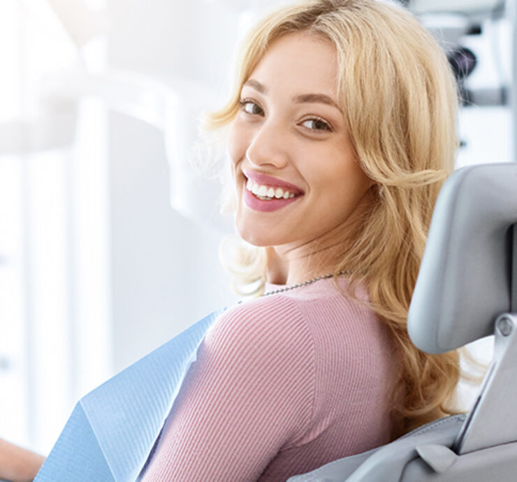 Smiling and relaxed young woman sitting at dental chair in modern clinic, waiting for her dentist. Happy attractive blonde lady attending luxury dental clinic